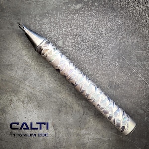 May include: A cylindrical titanium pen featuring a metallic finish with a repeating etched floral pattern across the barrel. The pen has a pointed tip and a smooth end cap. The text CALTI TITANIUM EDC is printed in dark blue in the lower left corner.