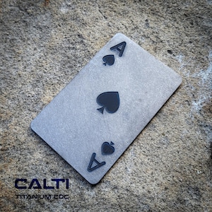 Puede incluir: Una carta de juego de titanio gris, el As de Picas. La carta presenta un símbolo de pica negro y la letra "A" en cada esquina. El texto "CALTI TITANIUM EDC" es visible en la parte inferior de la carta.