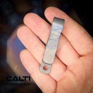 Puede incluir: Un pequeño clip metálico curvado con una superficie texturizada y detalles azules. El clip tiene un pequeño agujero en un extremo y el texto "CALTI TITANIUM EDC" en la parte inferior. El clip se sostiene en una mano.