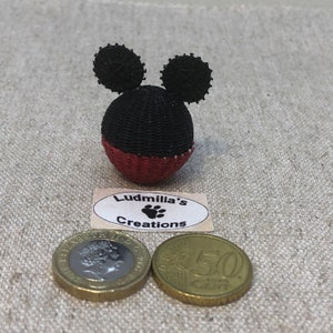 Puede incluir: Adorno artesanal con forma de Mickey Mouse. El adorno es negro y rojo, con dos orejas circulares negras. La base es roja y la cabeza es negra. Una etiqueta debajo dice "Ludmilla's Creations". Dos monedas están al lado del adorno.