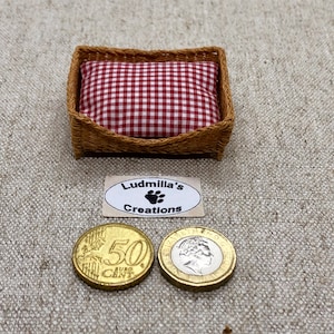 Op de afbeelding: Een klein dierenbedje van gevlochten wilg met een rood-wit geruit kussen. Het bedje is ongeveer zo groot als een 50 eurocent munt. Het label "Ludmilla's Creations" is zichtbaar onder het bedje.