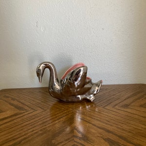 May include: A silver swan-shaped ceramic planter with a pink interior. The swan has a long, curved neck and is positioned on a wooden table with a geometric pattern. The planter is a decorative item.