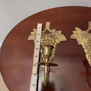 May include: A pair of gold-toned brass candle holders with a grapevine design. Each holder is approximately 6 inches tall.