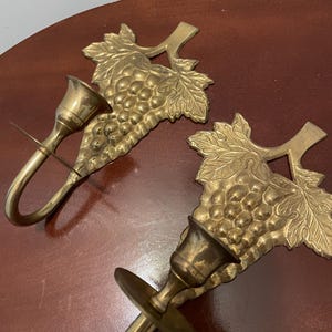 May include: A pair of brass wall sconces with a grapevine design. Each sconce has a single candle holder and a hook for hanging.