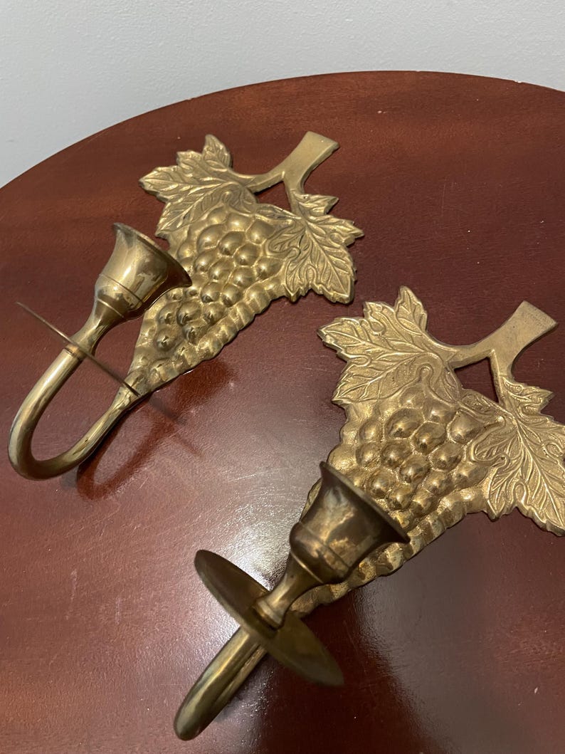 May include: A pair of brass wall sconces with a grapevine design. Each sconce has a single candle holder and a hook for hanging.