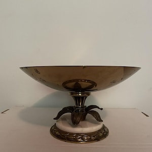 Puede incluir: Un cuenco decorativo de latón con una base ancha y poco profunda. El cuenco se asienta sobre un pedestal detallado con un diseño floral y una base de mármol blanco. El diseño es vintage y elegante.