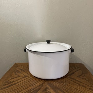 May include: White enamel stock pot with black rim and black knob on the lid. The pot is sitting on a wooden table.