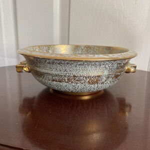 May include: A decorative bowl with a speckled blue-green and bronze exterior, accented with gold trim and handles. The bowl has a wide, shallow shape and sits on a small, gold-colored base. The interior is also gold.