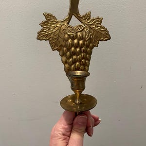 May include: A gold metal wall-mounted candle holder with a grapevine design. The holder has a single candle cup and a decorative grape cluster.