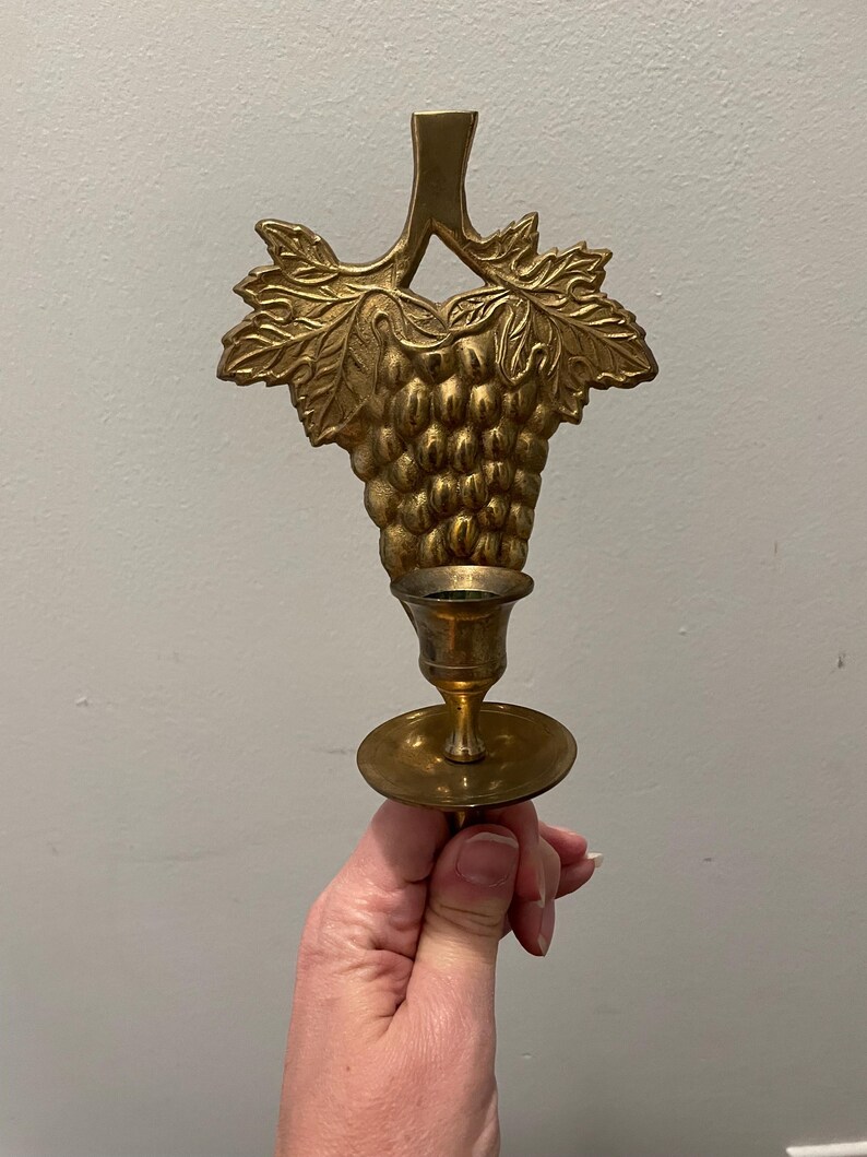 May include: A gold metal wall-mounted candle holder with a grapevine design. The holder has a single candle cup and a decorative grape cluster.