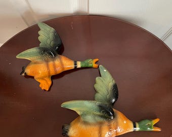 May include: Two ceramic duck figurines with orange, green, and black paint. The ducks are in mid-flight with their wings spread out. They are facing each other with their beaks open.