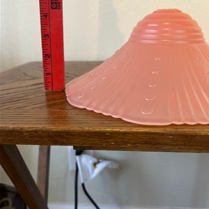 Peut inclure: Un abat-jour de lampe en verre rose avec un bord festonn&eacute; et un motif textur&eacute;. L'abat-jour est pos&eacute; sur une table en bois.