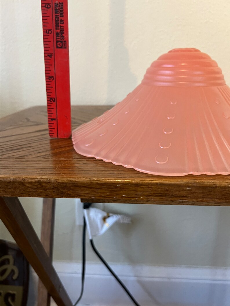 Peut inclure: Un abat-jour de lampe en verre rose avec un bord festonn&eacute; et un motif textur&eacute;. L'abat-jour est pos&eacute; sur une table en bois.