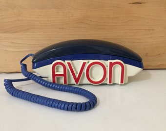 Könnte beinhalten: Ein weißes und blaues Vintage-Drehwahltelefon mit einem blauen Spiralkabel. Das Telefon hat das Wort "AVON" in roten Buchstaben auf der Vorderseite.