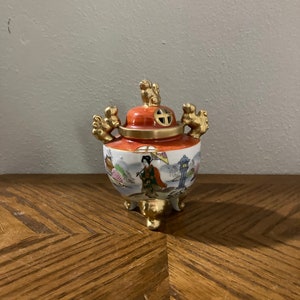 May include: A small, orange and white ceramic incense burner with gold accents. The burner features a scene of a woman in traditional Japanese clothing walking with an umbrella. The burner has three gold feet and a lid with a small hole in the center.