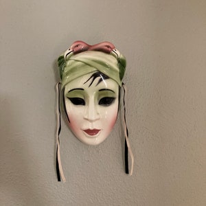 Könnte beinhalten: Eine weiße Keramikmaske mit einem grün-rosa Turban, schwarzen und weißen Bändern und einem bemalten Gesicht.