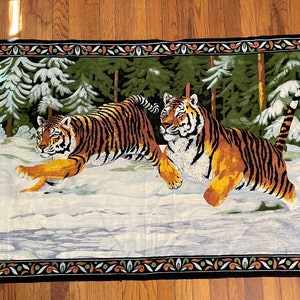 May include: A tapestry wall hanging featuring two tigers in a snowy forest setting. The tigers are depicted in a realistic style with orange and black stripes. The forest is green with snow-covered trees. The tapestry has a black border with a green and brown floral design.