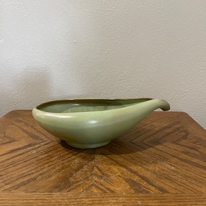 Puede incluir: Un macetero de cerámica verde claro con forma de lágrima y borde marrón. El macetero está sobre una mesa de madera.