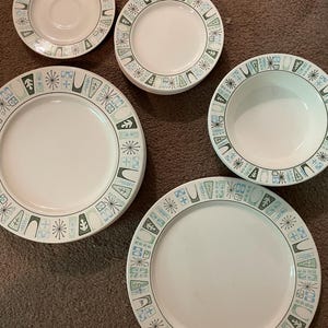 Peut inclure: Ensemble d'assiettes et de bols en céramique blanche avec une bordure décorative. La bordure présente un motif répétitif de formes géométriques dans des tons de bleu, vert et gris. Comprend une soucoupe, des bols et des assiettes.
