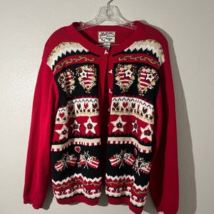 May include: Red knitted cardigan with a button-up front and a colorful pattern of hearts, stars, and flags. The cardigan has a white tag with the text "The Country Collection" on it.