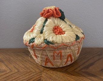 May include: A small, woven basket with a lid decorated with orange and green flowers. The basket is made of natural materials and has the word "AMA" woven into the side.