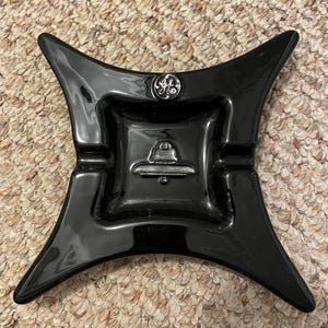 Może przedstawiać: Czarny popielniczkę ceramiczny z wzorem gwiazdy. Popielniczka ma logo GE i wypukły wzór w środku.