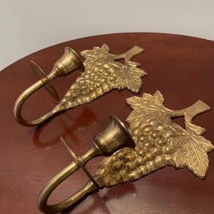 May include: A pair of brass wall sconces with a grapevine design. Each sconce has a candle holder and a hook for hanging.