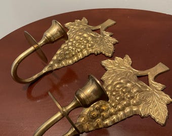 May include: A pair of brass wall sconces with a grapevine design. Each sconce has a candle holder and a hook for hanging.