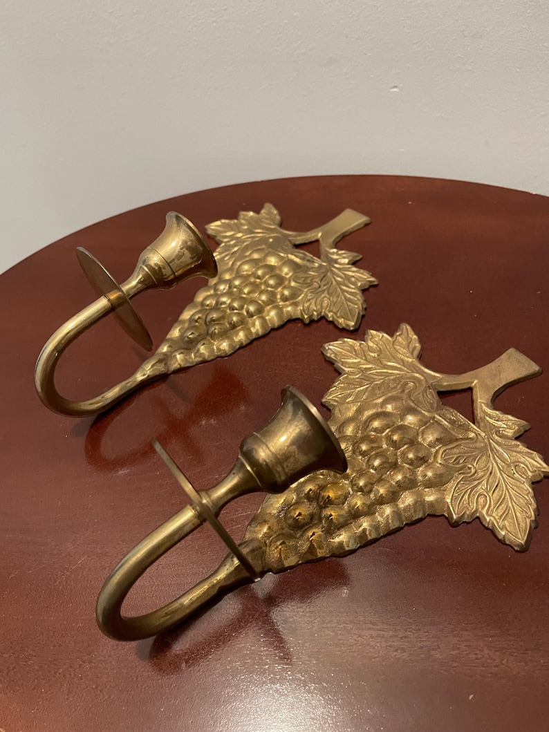 May include: A pair of brass wall sconces with a grapevine design. Each sconce has a candle holder and a hook for hanging.