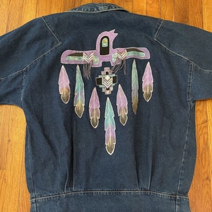 May include: Dark blue denim jacket with a colorful Native American-inspired design on the back. The design features a purple bird with feather accents in shades of purple, teal, and beige. The jacket has a classic collar and button closure.