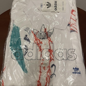 Op de afbeelding: Wit Adidas poloshirt met een kleurrijk abstract ontwerp en het Adidas logo. Het shirt zit in een plastic zak met een label waarop "Adidas Sports Textiles" staat.