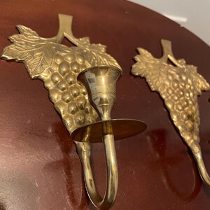 May include: A pair of gold-toned brass wall sconces with a grapevine design. Each sconce has a candle holder and a decorative grape cluster.
