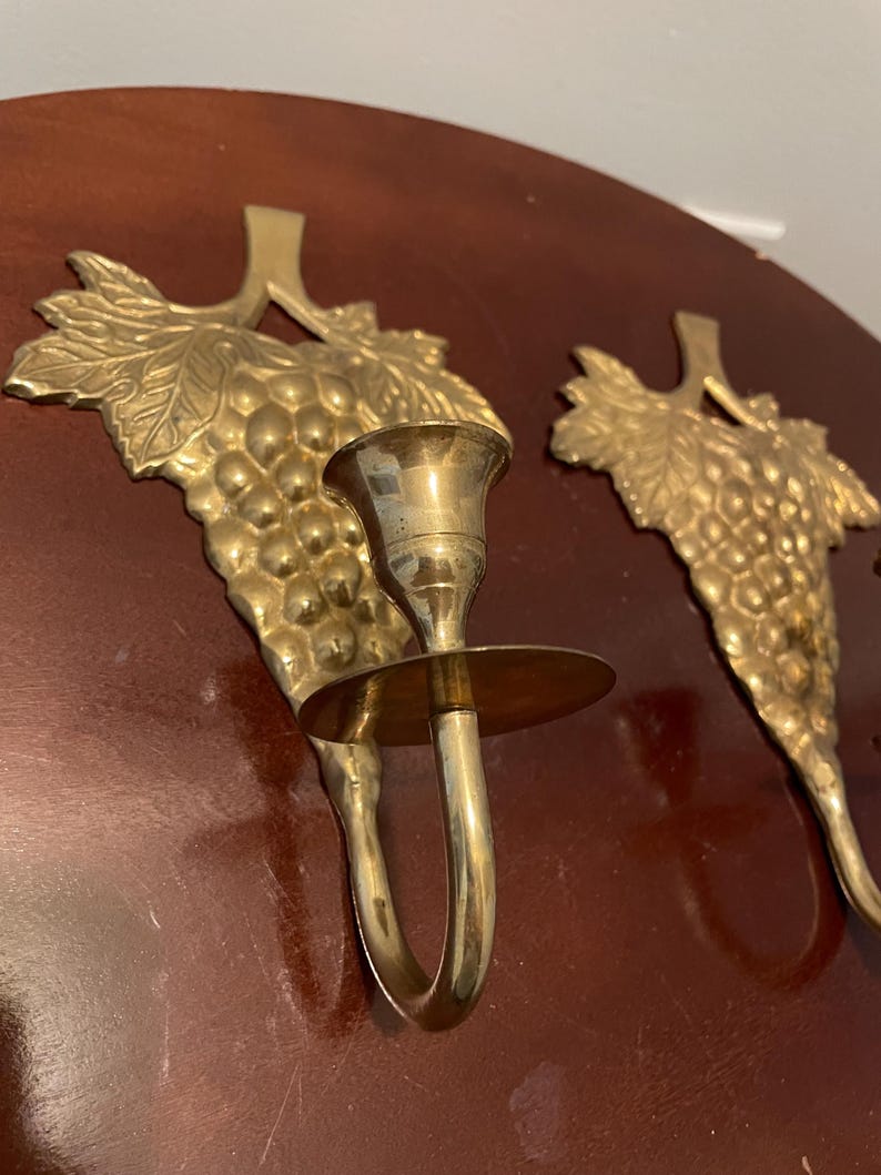 May include: A pair of gold-toned brass wall sconces with a grapevine design. Each sconce has a candle holder and a decorative grape cluster.