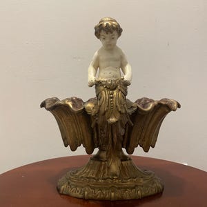 May include: A gold-toned metal decorative stand with a cherub figure on top. The stand has a scalloped edge and a detailed base.