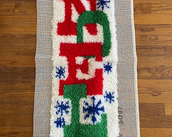 May include: A white canvas with a red, green, and blue yarn rug depicting the word "Noel" with snowflakes.