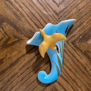 May include: A blue ceramic wall vase shaped like a flower with a yellow bird perched on top. The vase has brown painted details.