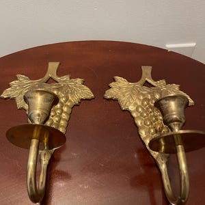 May include: A pair of gold-toned brass wall sconces with a grapevine design. Each sconce has a single candle holder and a hook for hanging.
