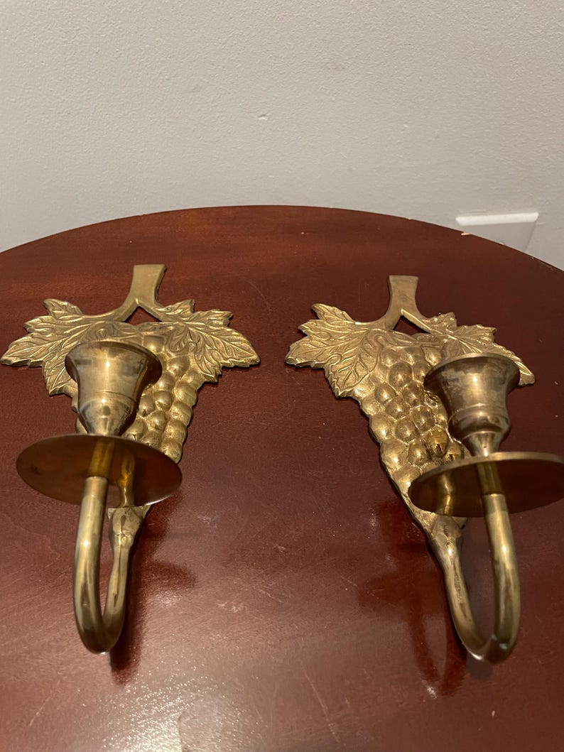 May include: A pair of gold-toned brass wall sconces with a grapevine design. Each sconce has a single candle holder and a hook for hanging.