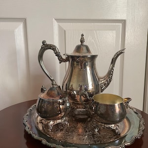 Puede incluir: Un juego de té plateado sobre una bandeja redonda. El juego incluye una tetera, un azucarero y una crema. La bandeja y todas las piezas tienen detalles ornamentados y se exhiben sobre una mesa de madera oscura.