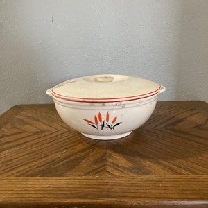 Puede incluir: Un tazón de servir de cerámica blanca con tapa y un diseño rojo y negro de juncos estilizados. El tazón está sobre una mesa de madera.