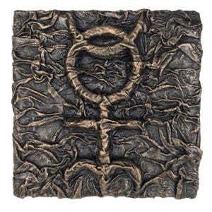 Puede incluir: Arte mural cuadrado y texturizado con un acabado metálico oscuro. Un símbolo de color bronce, que se asemeja a un círculo con una cruz debajo, está centrado. La obra de arte tiene una apariencia rugosa y arrugada, creando un diseño abstracto único.