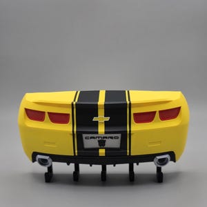 May include: A yellow toy car rear end with black racing stripes and the word "CAMARO" on the license plate. The car has red taillights and silver exhaust pipes. The background is a solid gray.