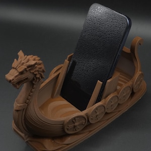 Könnte beinhalten: Ein brauner, 3D-gedruckter Telefonhalter in Form eines Wikinger-Langschiffs. Der Halter hat einen Drachenkopf am Bug und Schilde an den Seiten. Ein schwarzes Smartphone befindet sich im Inneren des Halters, der das Telefon aufrecht halten soll.