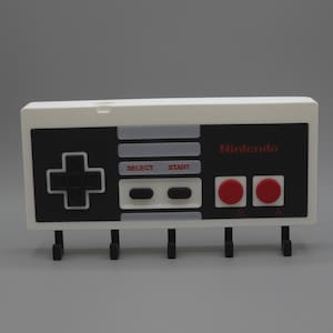 May include: A wall-mounted key holder in the shape of a Nintendo controller, coloured white and black. It features a directional pad, buttons, and the Nintendo logo. It has five black hooks for keys or small items.
