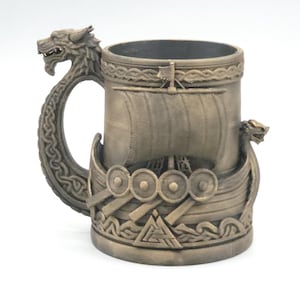 May include: A bronze-coloured mug featuring a relief of a Viking longship with a sail and shields. The handle is shaped like a dragon head with intricate knotwork patterns. A triangular symbol is embossed at the base of the ship on the mug exterior.