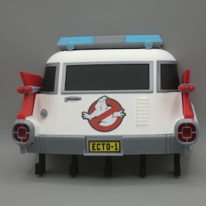 Puede incluir: Un llavero de pared blanco con forma de la parte trasera del vehículo Ecto-1. Presenta aletas traseras rojas, una barra de luces azul en el techo, ventanas negras, un logotipo de Ghostbusters y una matrícula amarilla con el texto ECTO-1. Cinco ganchos negros cuelgan debajo.