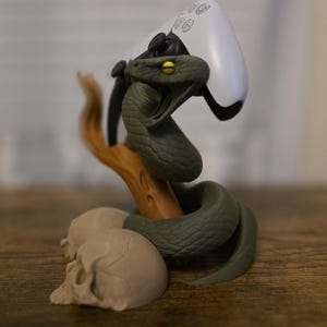May include: A green snake-themed gaming controller stand. The snake is coiled around a wooden branch, with a white controller resting on top. Two skulls are at the base of the stand. The snake has yellow eyes.