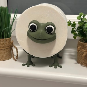 Könnte beinhalten: Ein skurriler, froschförmiger Toilettenpapierhalter in Grün, der eine Rolle weißes Toilettenpapier hält. Der Frosch hat große schwarze Augen und einen lächelnden Mund. Zwei kleine Topfpflanzen in Jutesäcken flankieren den Halter.