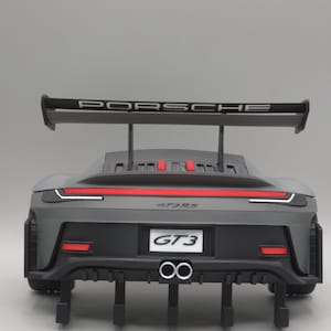 May include: A grey and black toy Porsche GT3 car, viewed from the rear. The car has a large rear wing with the word "PORSCHE" in white. Red detailing is present on the lights and bumper. Dual exhaust pipes are visible.