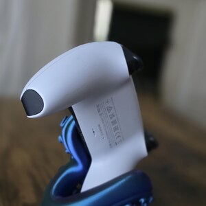 Puede incluir: Un mando de videojuegos blanco y negro apoyado en un soporte iridiscente azul y morado. El soporte tiene un diseño único y estilizado. El mando está ligeramente inclinado, mostrando su forma y el soporte.